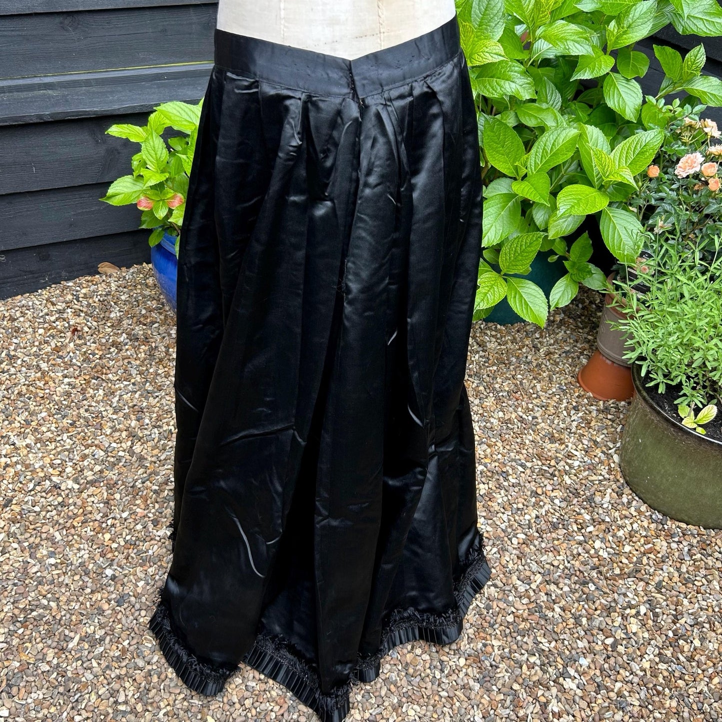 Antique black sateen walking skirt with finely pleated hem set against a shingle and green planted background