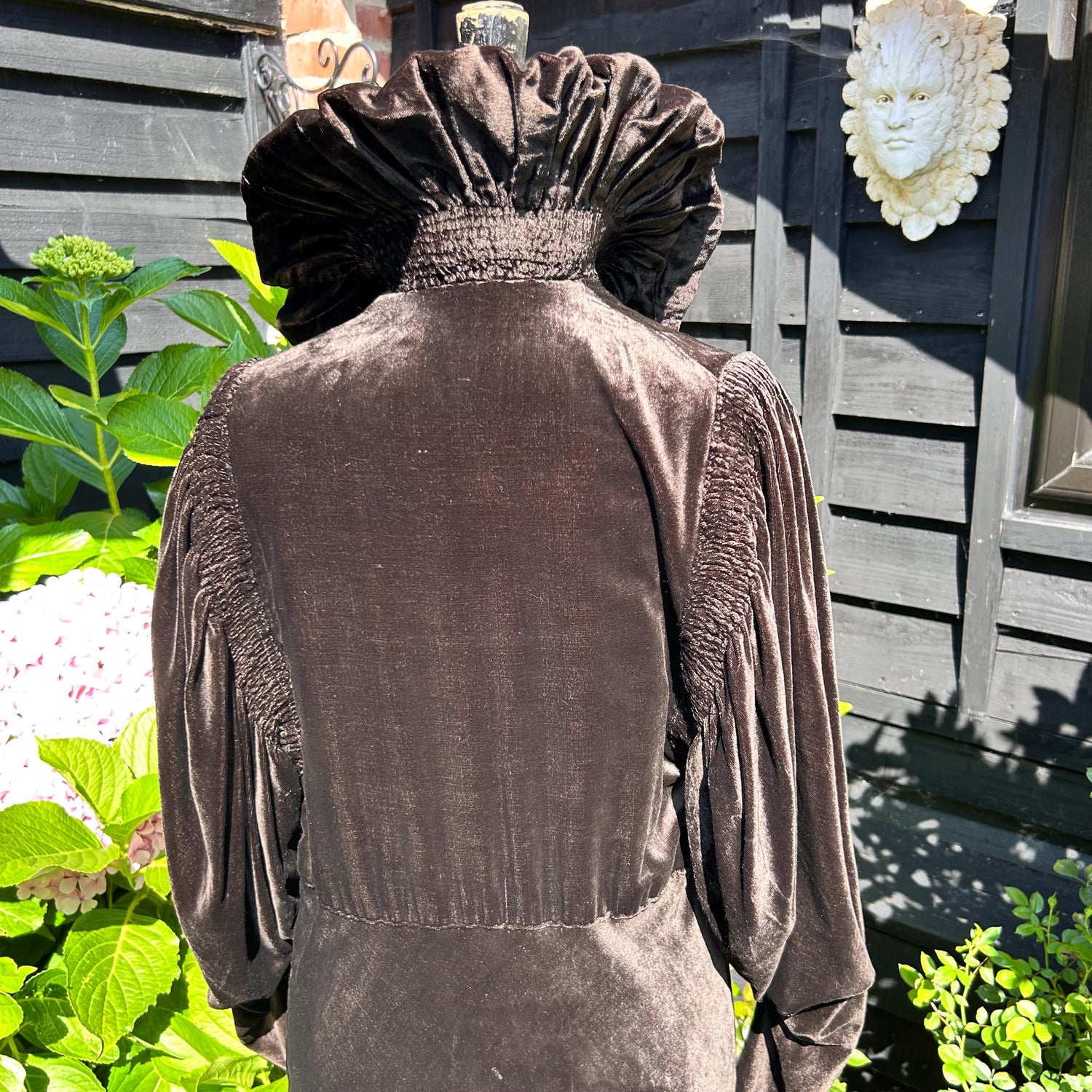 Back view of 1930s clothing, a brown velvet coat with upturned feature collar in front of green plants & black fence