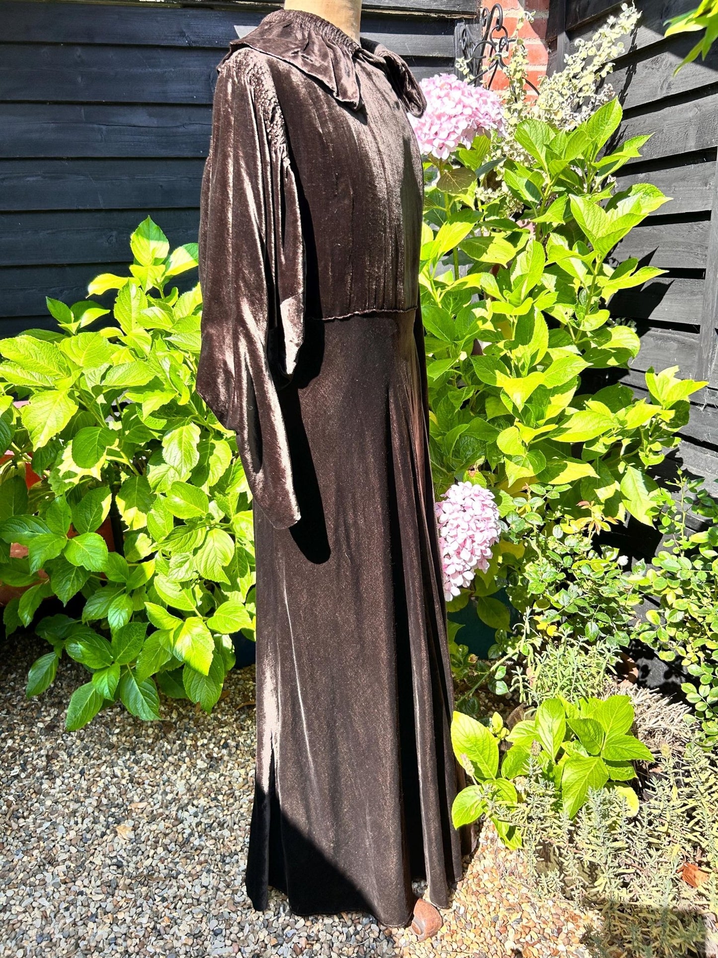 Dark Brown long Velvet Coat with Smocked Sleeves side view against green plants black fence & stone - Splendid Vintage World