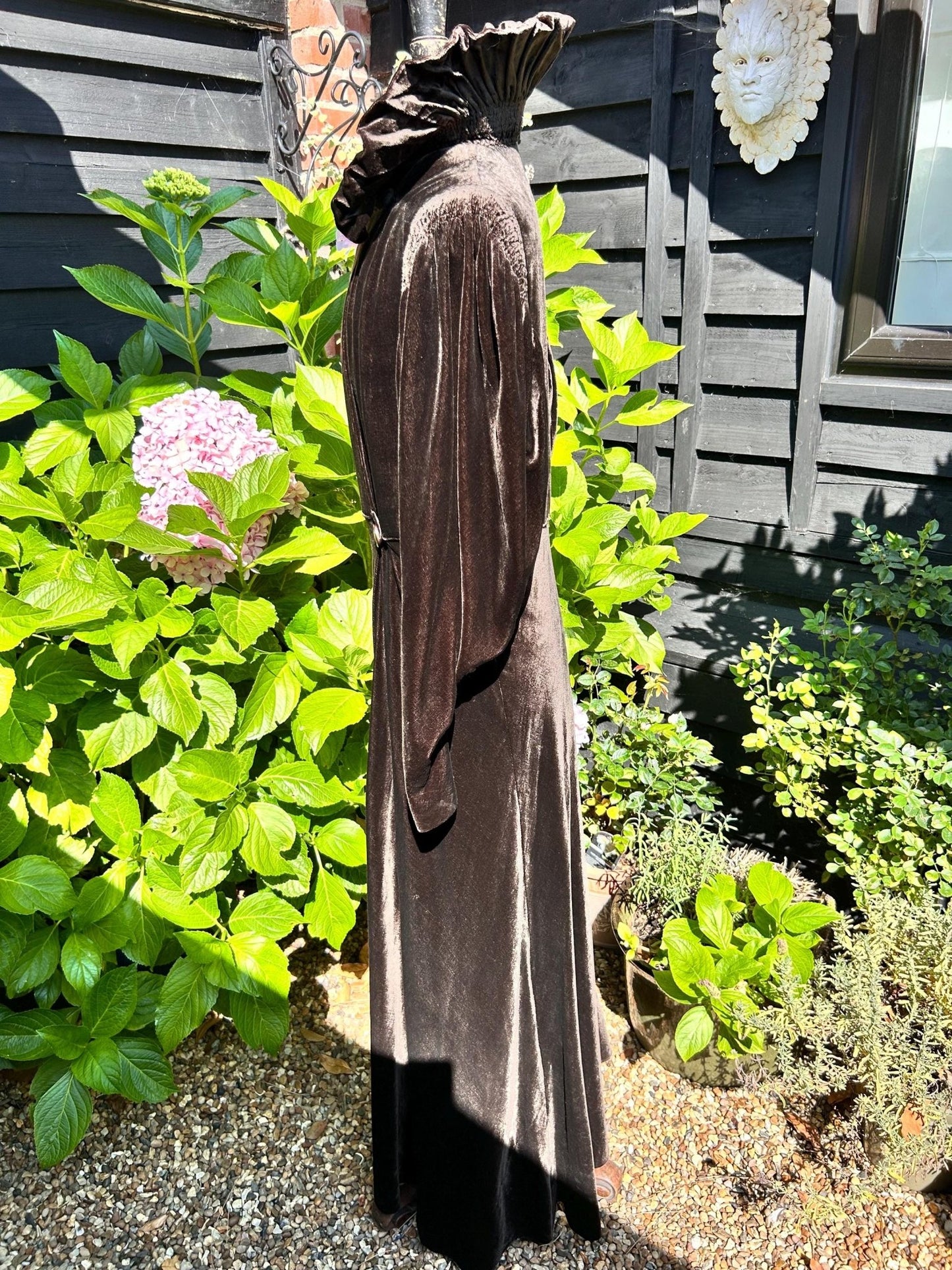 side view of Brown long Velvet Coat, upturned collar Smocked Sleeves against plants & black fence- Splendid Vintage World