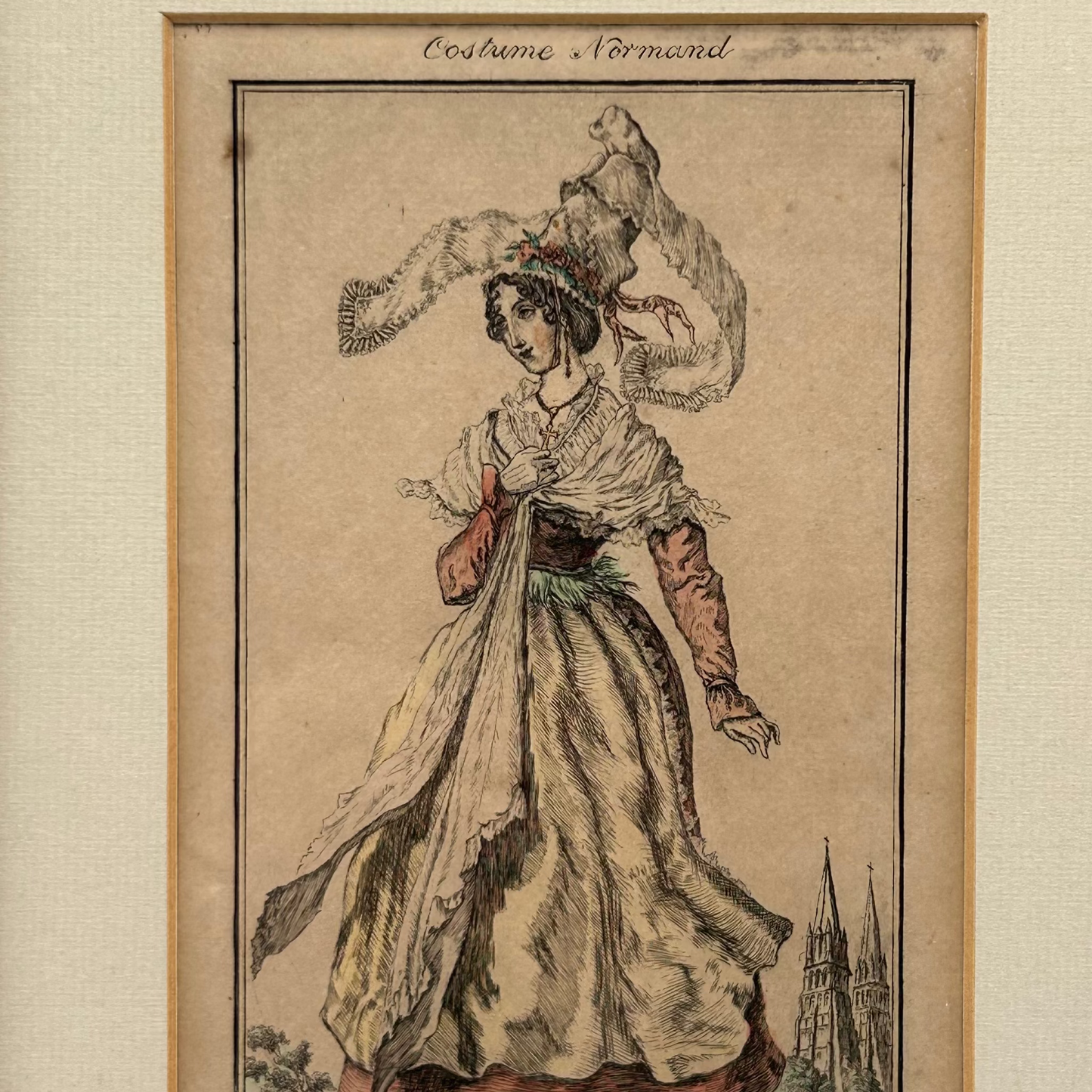 Antique print of a woman in Normand costume in tall hat on a beige paper featuring a church with 2 spires in the distance