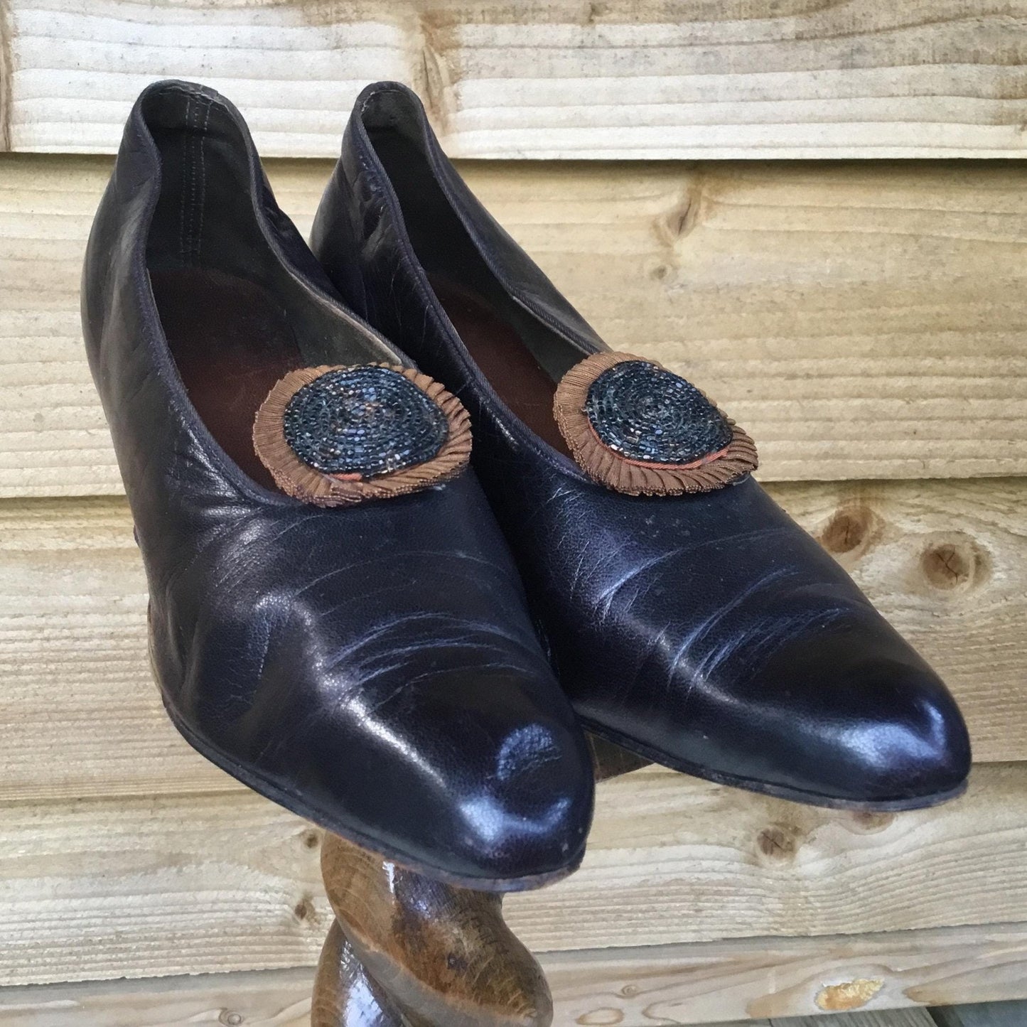 Antique 1910s deep purple Leather Shoes with beaded rosettes on wooden stand against fence backdrop - Splendid Vintage World