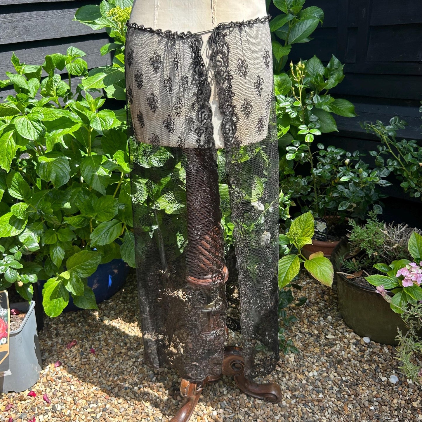 Antique black lace bonnet veil, lace panel attached to a mannequin stood on gravel with green bushes and black fence behind