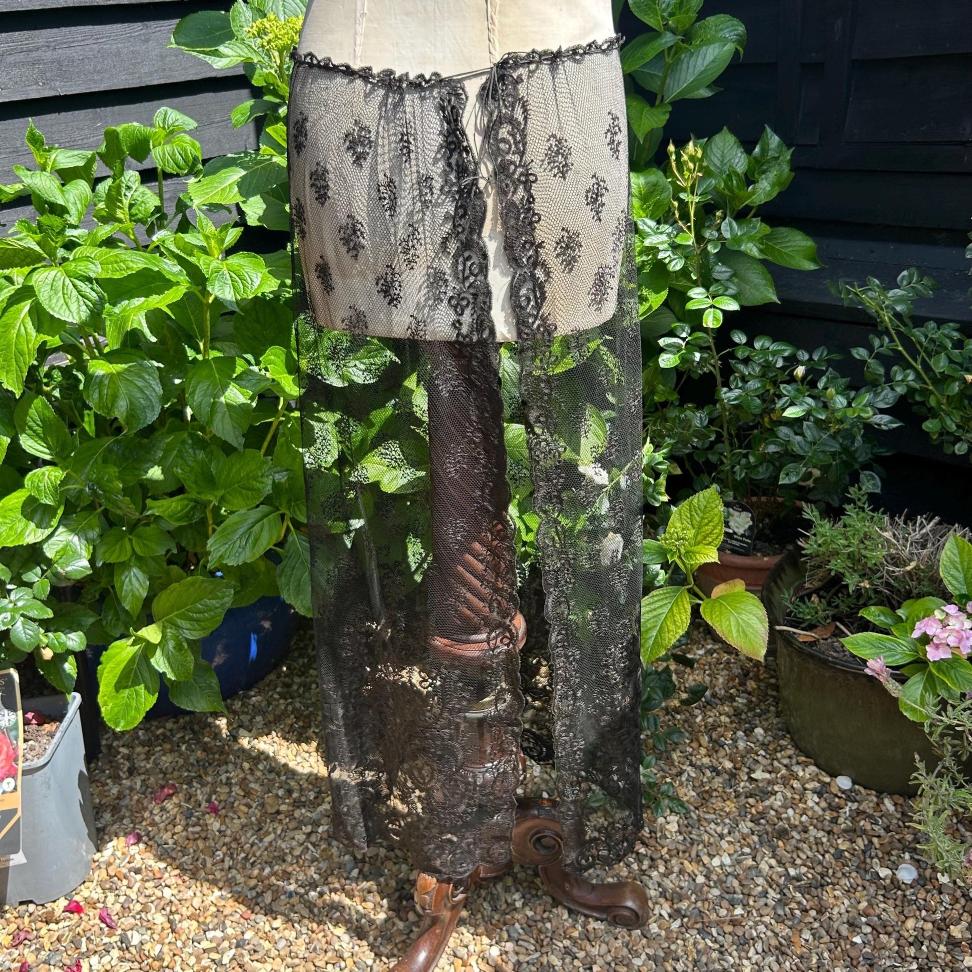 Antique black lace bonnet veil, lace panel attached to a mannequin stood on gravel with green bushes and black fence behind