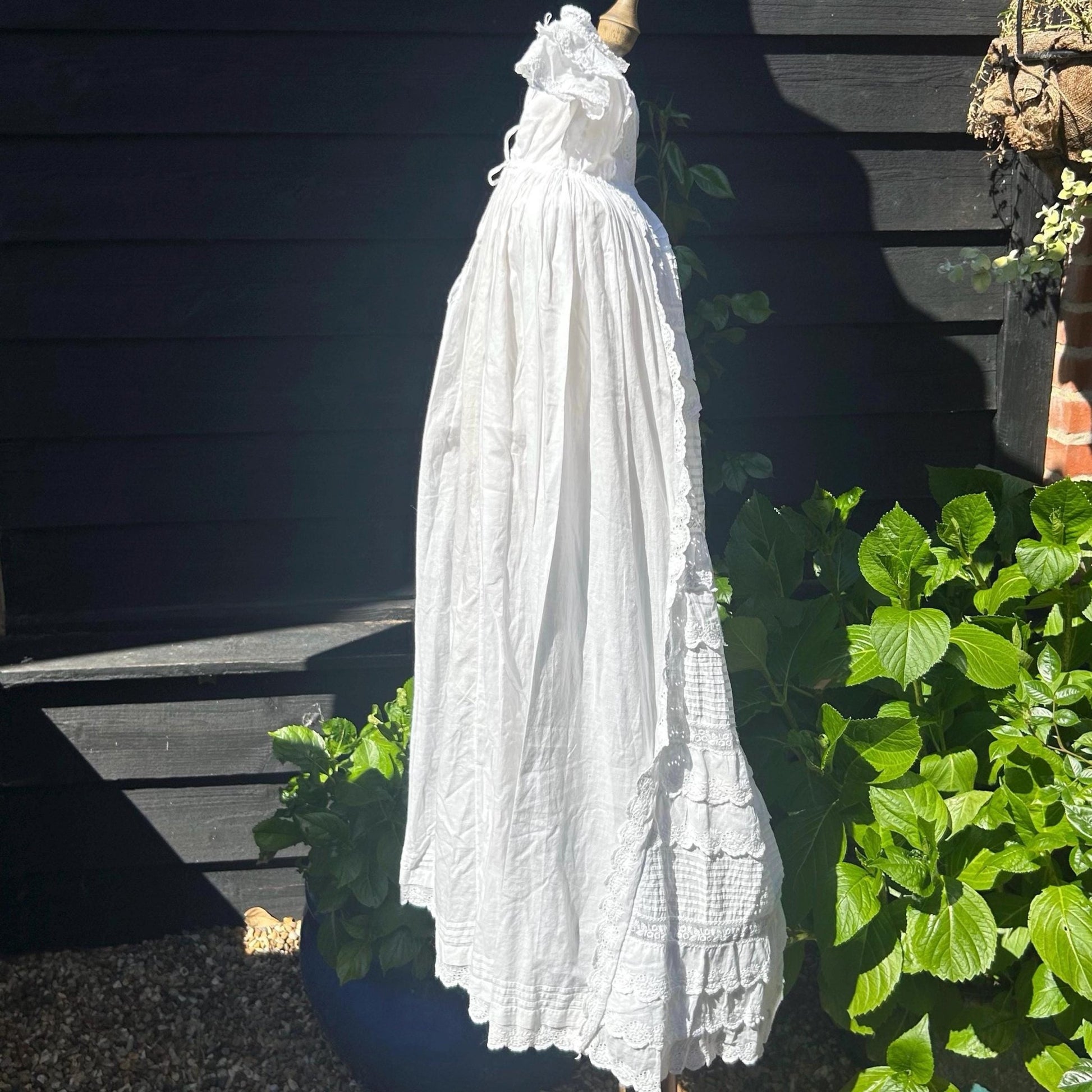 side view of white long Antique Christening Gown against black fence and greenery - Splendid Vintage World