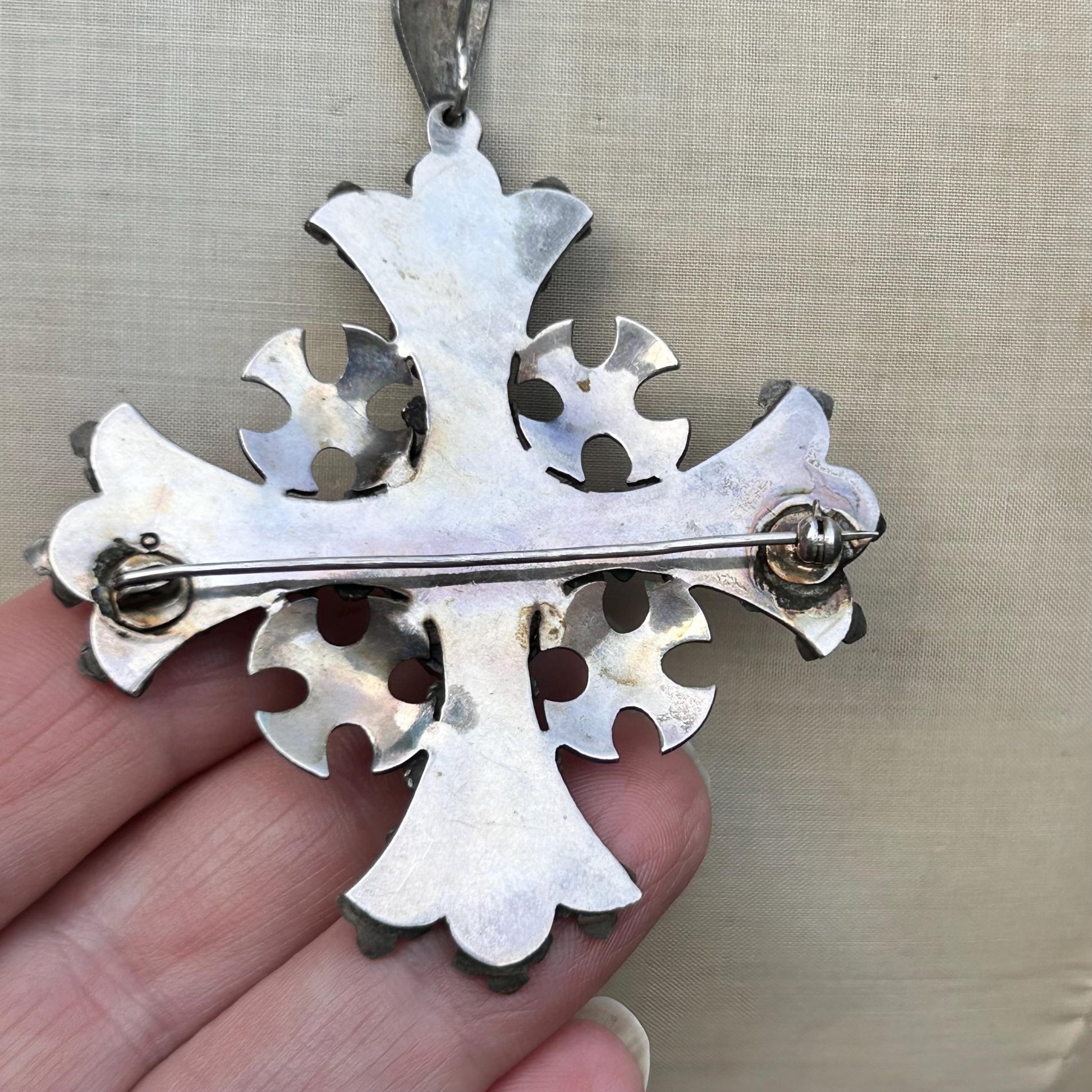back of silver cross pendant with brooch fitting held by hand on beige backdrop - Splendid Vintage World