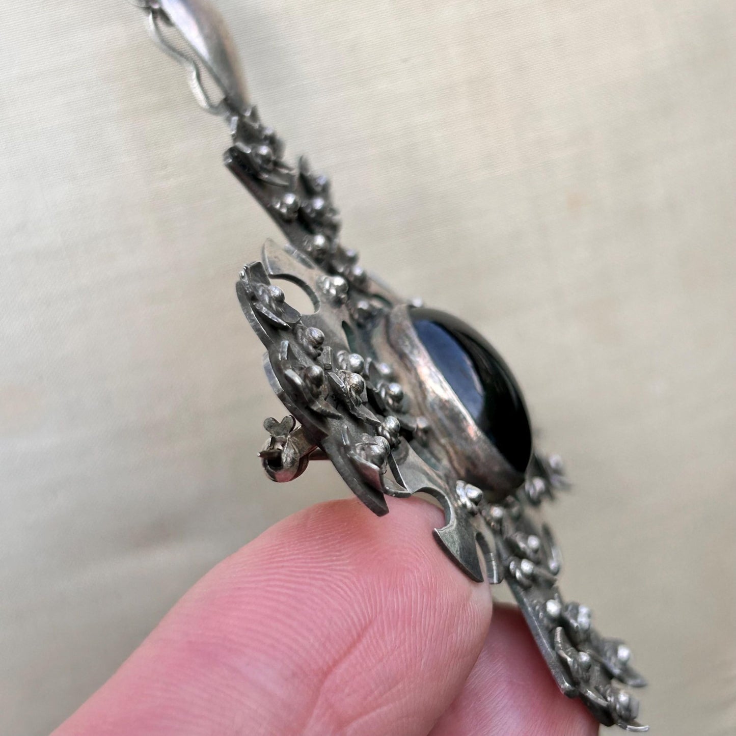 side view of silver cross pendant Set with black cabochon on silver chain held by hand on beige back - Splendid Vintage World