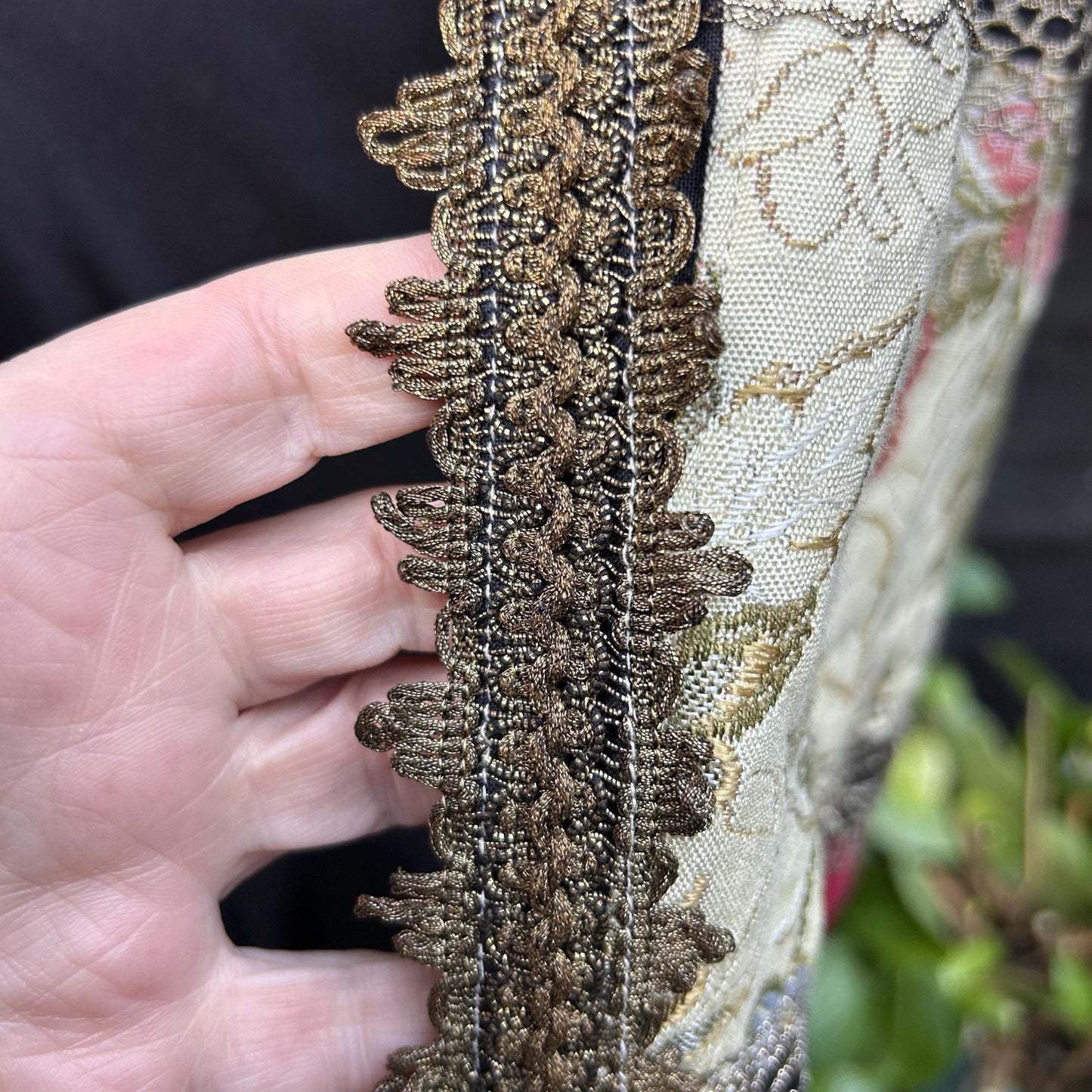 wide Gold Metallic Trim with patterned beige fabric held by hand with black and greenery background - Splendid Vintage World