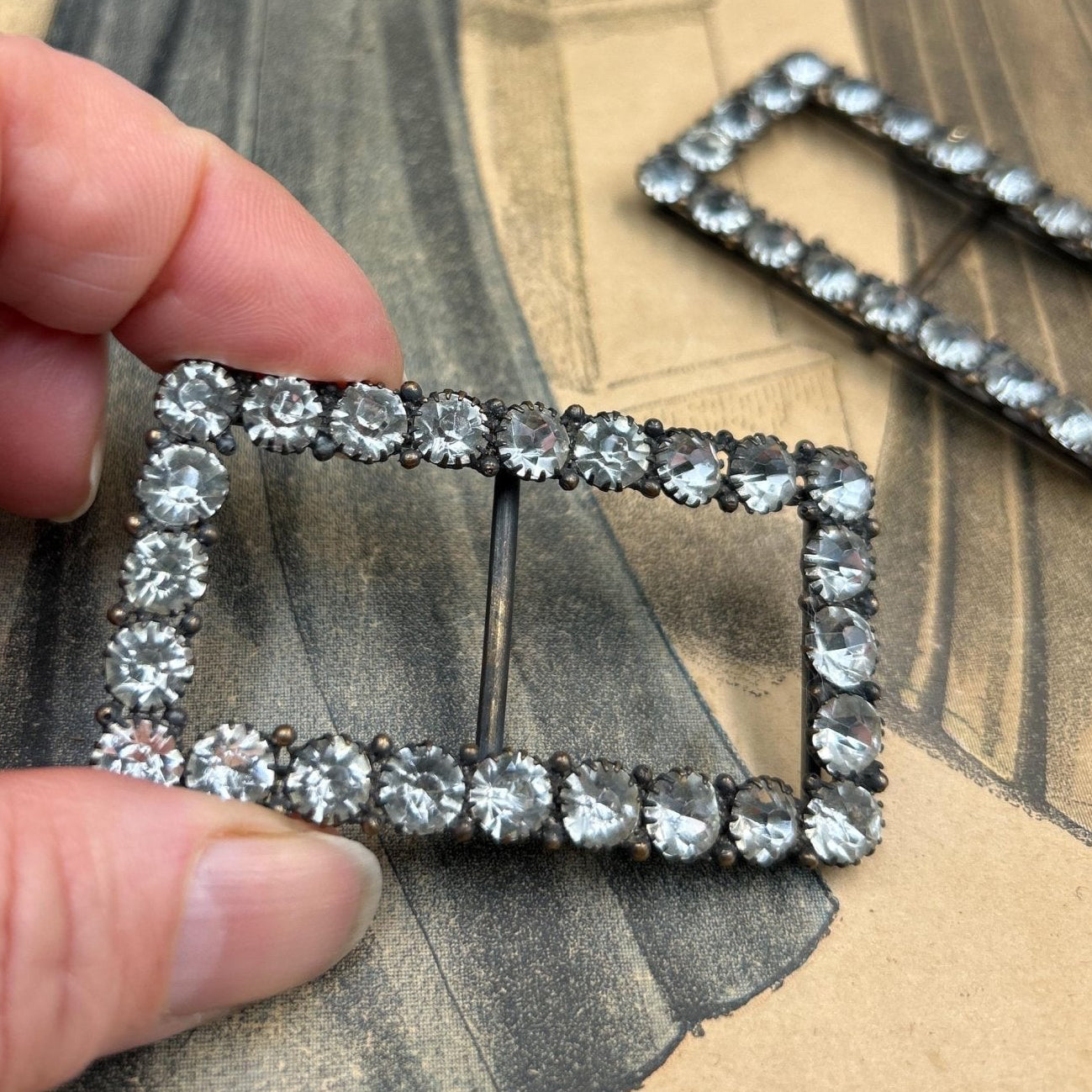2 Vintage sash buckles paste diamante 1 being held by 2 fingers against printed paper - Splendid Vintage World